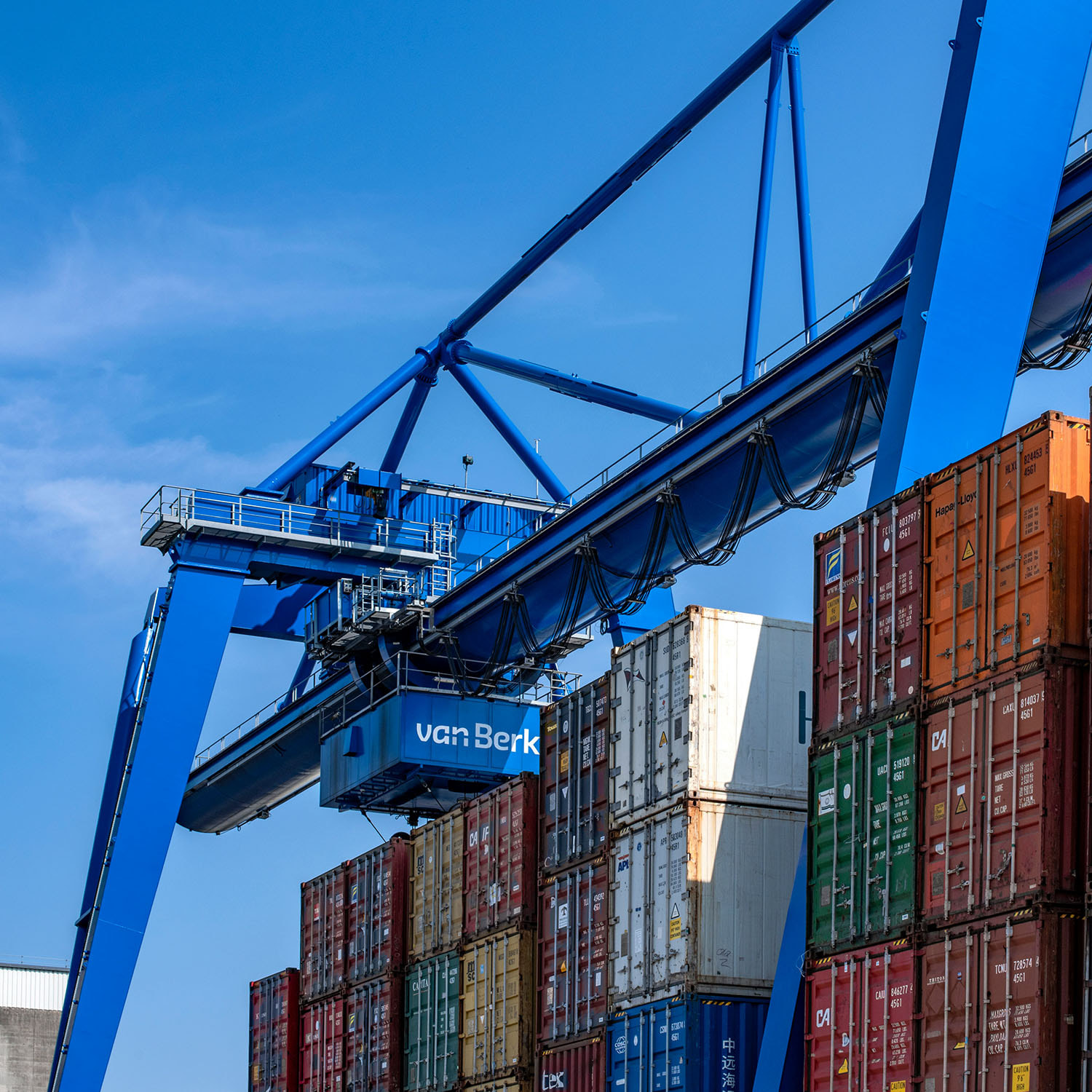 Ein großer, leuchtend blauer Containerkran der Marke van Berk hebt einen Frachtcontainer über mehrere Stapel bunter Schiffscontainer in einem Hafen unter klarem blauem Himmel.